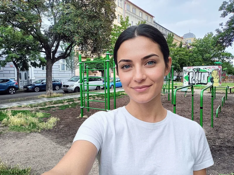 Jsem v bílém tričku před zelenými workoutovými konstrukcemi; za mnou jsou stromy, domy a nízká zeď s graffiti.